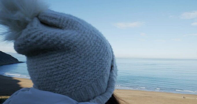 Mujer con gorro y chaqueta de abrigo desde la sombra mira al mar en los primeros d&iacute;as de primavera en un d&iacute;a soleado, plano desde atr&aacute;s mas corto.