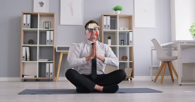 Portrait of funny tired business man employee meditating in office sitting on the floor practicing yoga on mat in Lotus pose with a notes on his face resting from a work. 4k video.