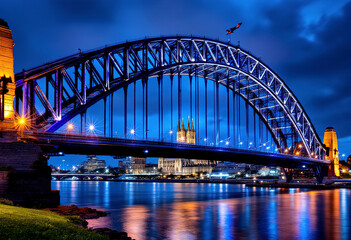 Obraz premium A large illuminated bridge with arched steel structures spanning a river at night, with a cathedral-like building visible in the background