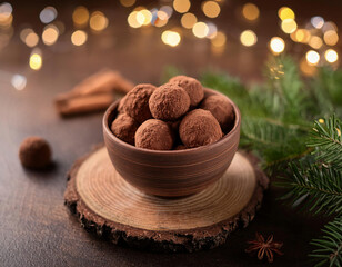 Chocolate truffles dusted with cocoa powder are served in a rustic brown cup surrounded by pine branches, pinecones, and twinkling holiday lights.