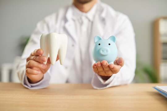 Portrait of a male dentist doctor sitting at the desk in dentistry clinic and holding piggy bank and healthy white tooth in hands. The concept of money saving for expensive dental treatment