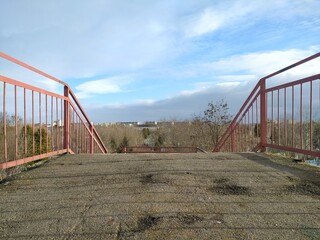 Red metal bridge over the river, with a view of the city. Metal handrails on the stairs in the park