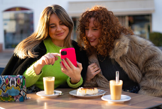 two very happy girls drinking coffee in a restaurant while looking at the cell phone smiling