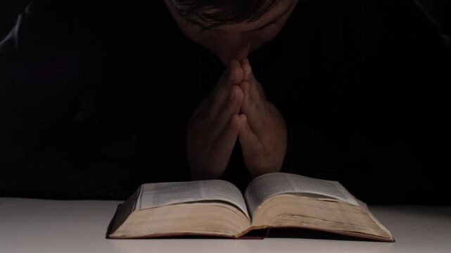 Priest praying with hands joined over open bible in dark room
