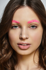 A confident young woman showcases her beauty with vibrant pink eyebrows against a grey backdrop.