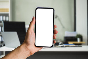 Hand holding white mobile phone with blank white screen in office.