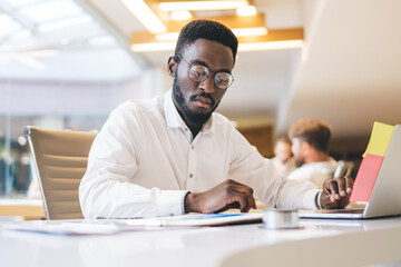 Fototapeta premium Concentrated african american male employee in eyewear for vision correction reading finance report and analyzing, serious dark skinned male manager sitting at working table with laptop computer