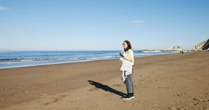 Mujer feliz se pone las gafas de sol, con gorro y chaqueta de abrigo en la mano en la playa al en los primeros d&iacute;as de primavera en un d&iacute;a soleado,  frente al mar. Plano general.