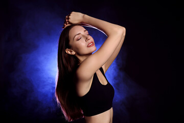 Beautiful woman in stylish sportswear on dark background with color light and smoke