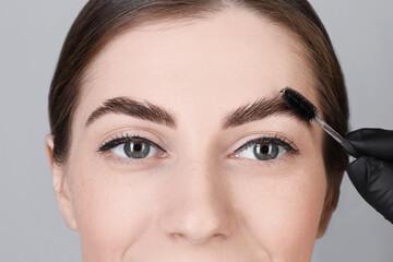 Brow lamination. Cosmetologist combing woman's eyebrows with brush against grey background, closeup