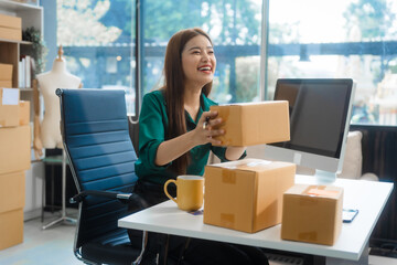 An Asian young woman sits in a room, working on a computer and making calls or video calls, managing her small business and online sales, surrounded by cardboard boxes ready for shipping.