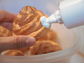 Female hand holds a round fancy choux pastry or eclair and fills it with white custard with the help of a confectionery syringe
