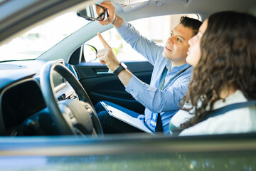 Driving instructor demonstrating safety procedures and adjusting rearview mirror for teen student during driving lesson