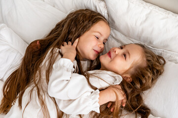 Cheerful little girls, two twins sisters in white dresses in bed. Children lying in bed and hugging each other. Good morning. textiles for the nursery. Have a nice day. High quality photo