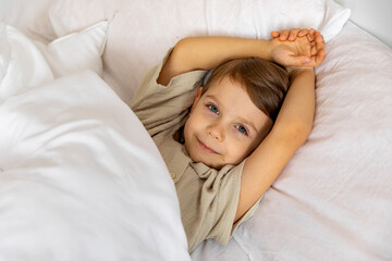 Cheerful little boy lying under white blanket in bed. Child woke up with a good mood in the morning. textiles for the nursery. Have a nice day. Motherhood concept. High quality photo. Copy space