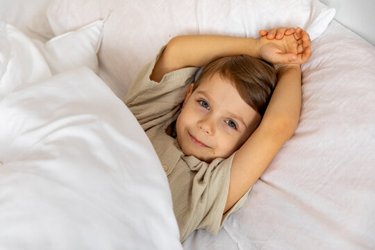 Cheerful little boy lying under white blanket in bed. Child woke up with a good mood in the morning. textiles for the nursery. Have a nice day. Motherhood concept. High quality photo. Copy space