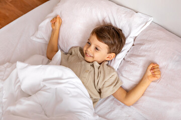 Cheerful little boy lying under white blanket in bed. Child woke up with a good mood in the morning. textiles for the nursery. Have a nice day. Motherhood concept. High quality photo. Copy space