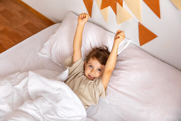 Cheerful little boy lying under white blanket in bed. Child woke up with a good mood in the morning. textiles for the nursery. Have a nice day. Motherhood concept. High quality photo. Copy space