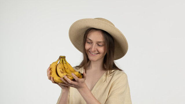 A woman dressed in a light outfit and straw hat joyfully examines a bunch of ripe bananas in a well-lit environment, showcasing natural beauty and freshness