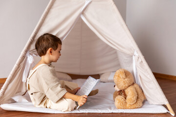 Little boy, 4-5 years old, sitting in a play wigwam tent, reading book with soft teddy bear. Little lover of reading books, interesting childhood, inquisitive child. Children room. High quality photo © Irina Mikhailichenko