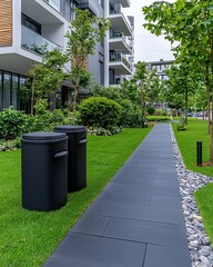 eco-conscious recycling natural resources innovation concept, A clean, landscaped walkway with green grass and trees, featuring two black trash bins beside a modern building.