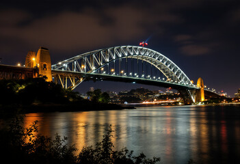 A large illuminated bridge arching over a body of water at night, with the bridge's reflection visible in the calm waters below