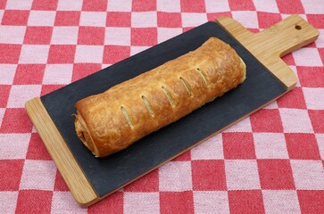 Sausage roll on a slate serving board lies on a red and white checkered tablecloth. Traditional delicacy for Lost Monday.