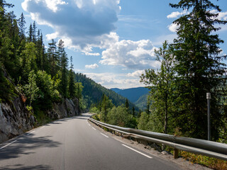 Fototapeta premium Scenic mountain road in Norway winding through lush greenery and under vibrant sky with scattered clouds.