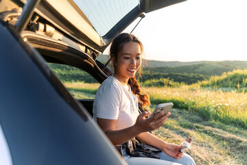 a young woman is sitting in the trunk of her car, having refreshments and checking her phone after a long hiking trip © DusanJelicic