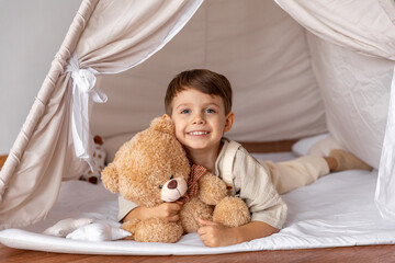 Happy 5 years old little boy lying in a wigwam tent, hugging soft toy teddy bear. Children's room,...