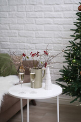 Glasses of champagne with Christmas dry tree branches with blurred lights and red berries, christmas tree on table in living room.