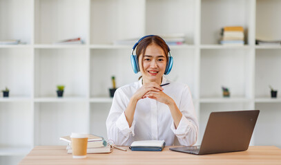 Concentrated young asian female student engaged in academic research with laptop and headphones at modern library desk, writing Notes and Studying Online.