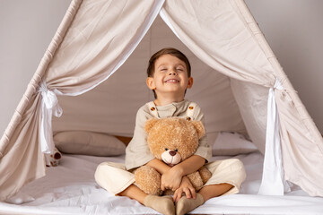 Happy 5 years old little boy sitting in a wigwam tent, hugging soft toy teddy bear. Children's room, childhood, games, imagination. High quality photo © Irina Mikhailichenko