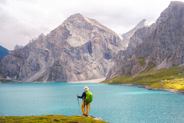 A woman stands on a rock with lush green grass by the Tientang Hu lake, Xinjiang, China. There are many mountains behind. The soft cool light shines on the still and shimmering blue water surface.