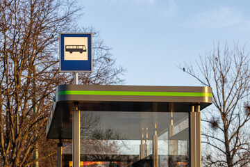 Bus stop sign on a modern shelter with green accents against a background of bare trees and clear...