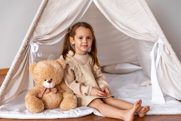 Happy 4-5 years old little girl sitting in a wigwam tent, hugging soft toy teddy bear. Children's room, childhood, games, imagination. High quality photo © Irina Mikhailichenko