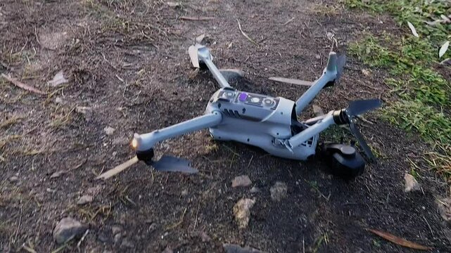 A crashed drone lies on the ground with a broken arm and a dislodged camera gimbal. Damaged propellers and blinking LEDs indicate its recent collision.