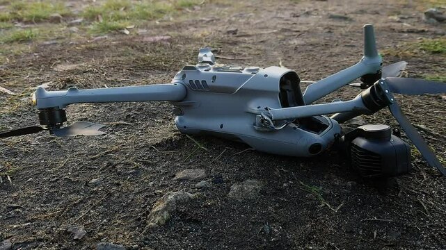 A damaged drone sits on the ground, broken propellers and a detached camera gimbal highlighting the result of a collision. Its LEDs blink faintly.