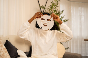 Young woman relaxing on sofa, applying hydrating face mask during peaceful skincare routine,...