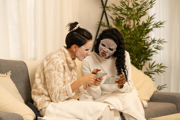 Diverse women sitting together on sofa, wearing protective face masks, reading application instructions during home skincare session