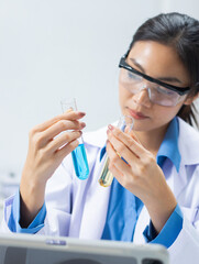 Professional Asian female chemist compares two liquid samples in lab, holding test tubes in both hands and studying, observing, analyzing result for medical and pharmaceutical research in medical lab.