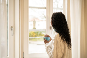Woman relaxing in bathrobe, sipping coffee near window, soft morning light illuminating tranquil...
