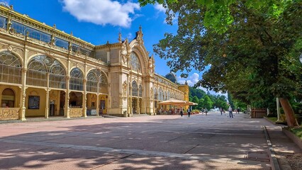 Fototapeta premium The Maxim Gorky Colonnade in Mariánske Lazne, Czech Republic with ornate architecture, large arched windows, and intricate details, surrounded by greenery and a bright blue sky.