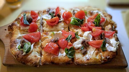 A freshly baked flatbread topped with cherry tomatoes, basil, mozzarella, olives, and anchovies, served on a wooden board, showcasing vibrant Italian flavors.