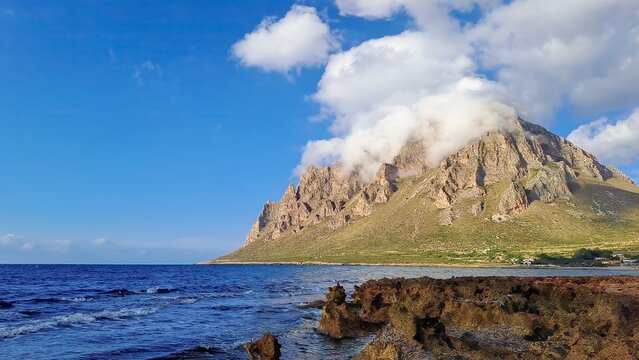 Baia di Cornino, Italy - A rugged mountain peak partially covered in clouds, with a rocky shoreline and calm blue sea under a clear sky, showcasing natural beauty.