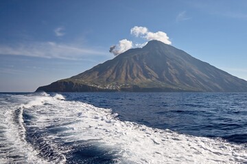 A striking view of Stromboli volcano rising from the blue sea, captured from a moving boat. The volcanic peak emits smoke under a clear sky, emphasizing the island's rugged terrain and natural power.