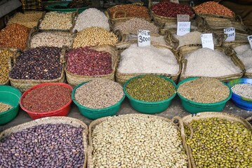 A bustling market stall showcasing a variety of pulses, beans, and rice in woven baskets and plastic bowls, with prices written on signs.
