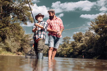 Two men friends fisherman fishing on river. Old father and son with rod fishing at riverside....