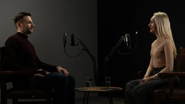 Man host and woman guest recording interview in stylish podcast studio using microphones, creating digital content, sit in chairs greet each other and start conversation.