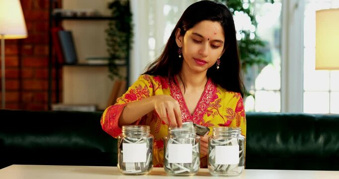 Confident Indian Asian young girl or wife in her mid-twenties, dressed in a kurta, placing Indian currency notes into three different glass jars, each with labels, for managing finances at home